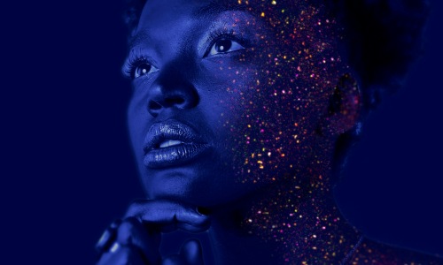 woman in a dark room with blue lighting and glowing fluorescent paint docs all over her face that glow like stars.